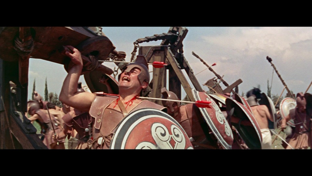 A chaotic battle scene featuring warriors in ancient armor, with one soldier reacting dramatically as he is struck by an arrow while surrounded by others holding shields.