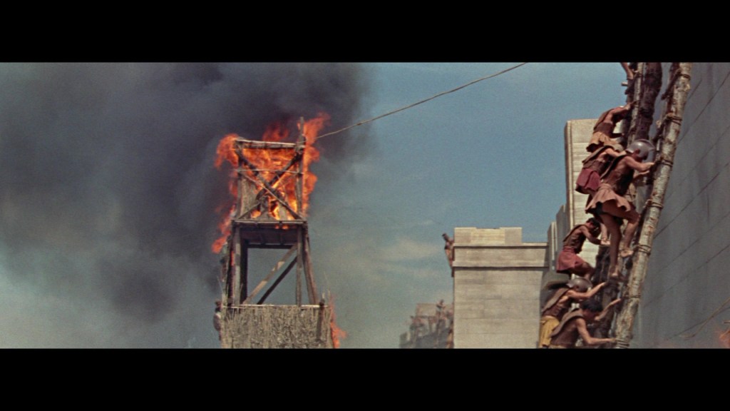 A wooden watchtower engulfed in flames amidst a dark smoke-filled sky, with soldiers climbing a ladder on a stone wall in the foreground.
