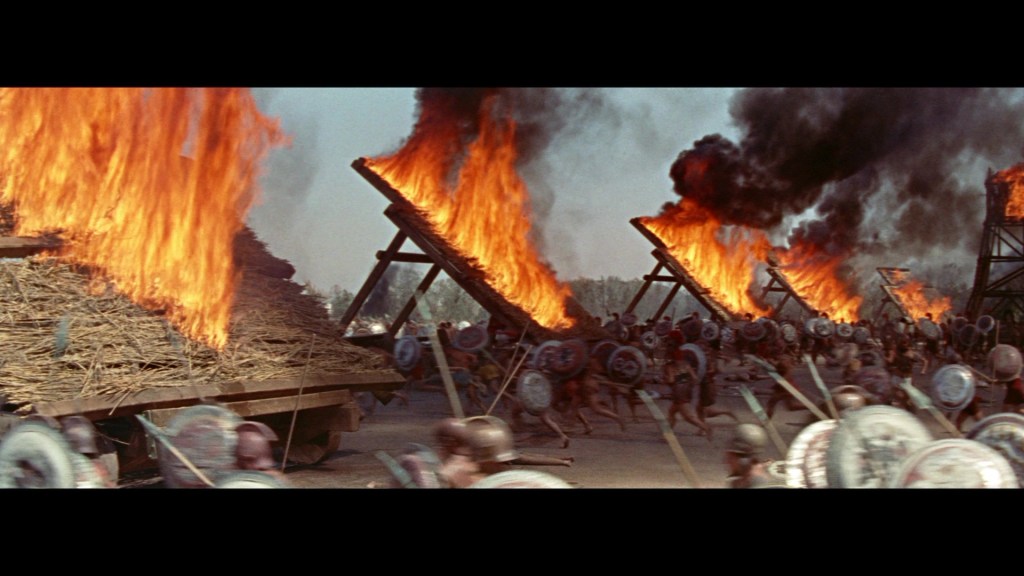 A scene depicting warriors in battle amidst large fires and smoke from burning structures, with soldiers charging forward, equipped with shields and spears.