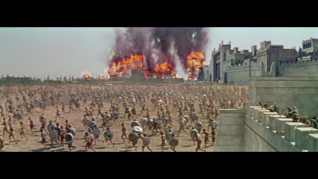 A large group of warriors in armor running on a battlefield, with structures burning and smoke rising in the background.