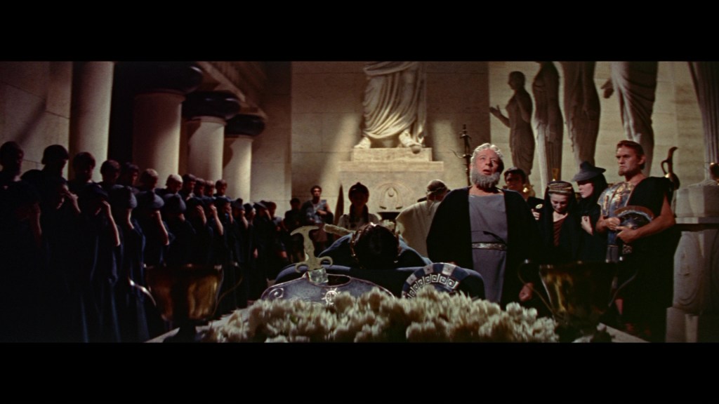 A dramatic scene depicting a gathering in an ancient setting, featuring a central figure standing and speaking, surrounded by a group of people dressed in historical attire, with sculptures and columns in the background.