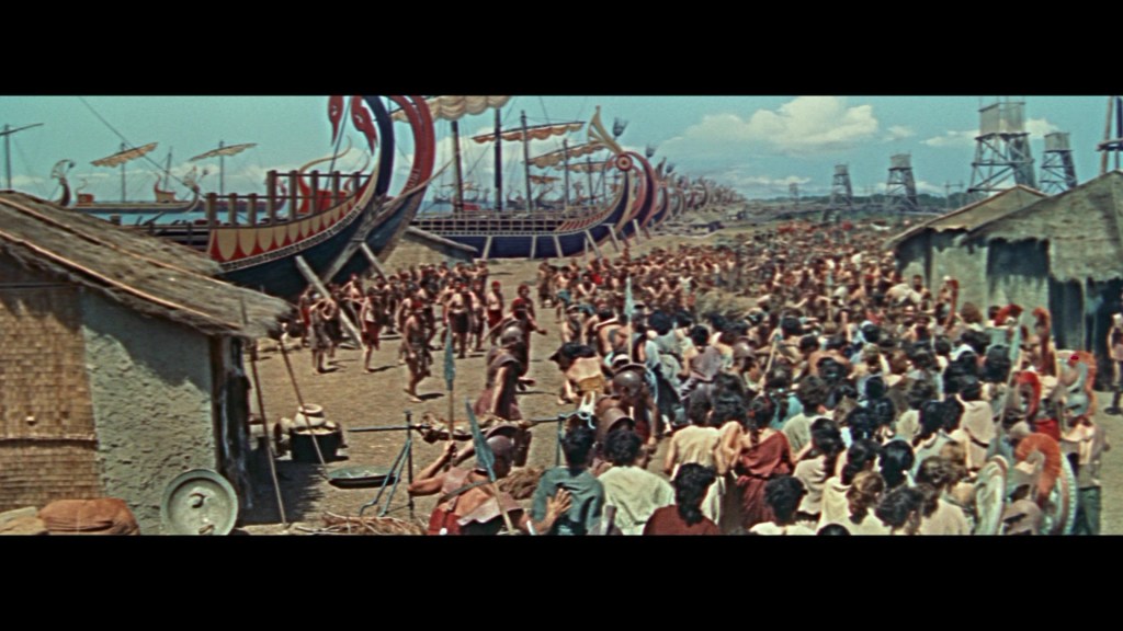 A crowd of people gather at a port adorned with large Viking ships, with thatched-roof huts in the foreground under a bright blue sky.
