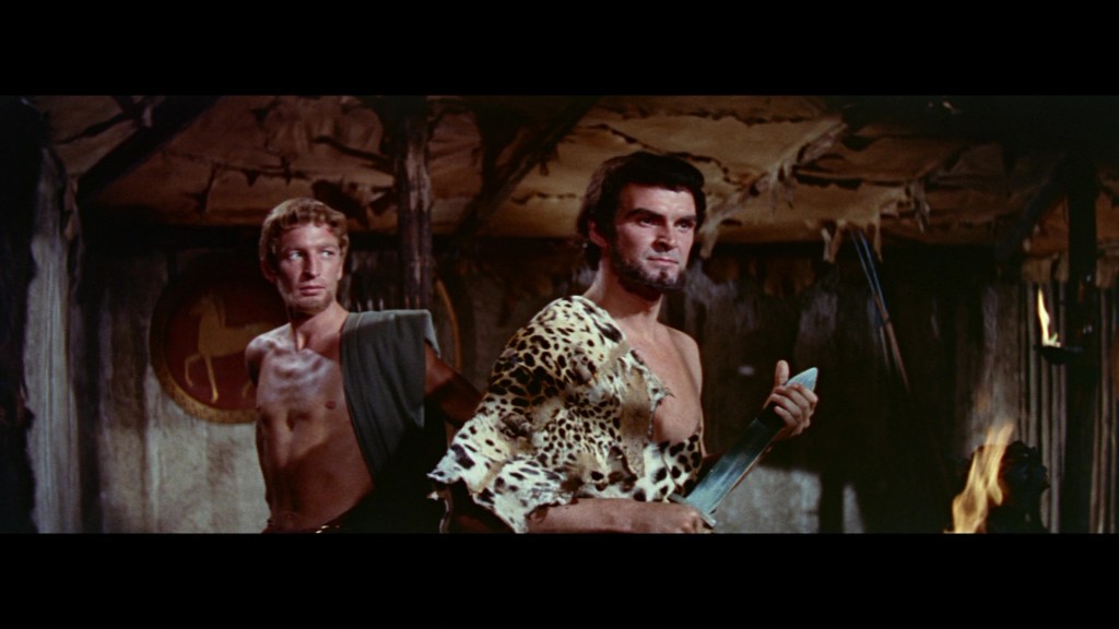Two men in prehistoric attire, one in a leopard-patterned tunic with a weapon, the other wearing a simple garment, standing in a rustic setting with a fire and wooden structures.