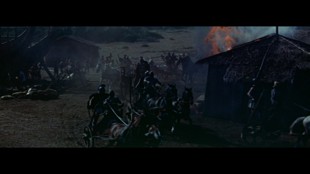 A chaotic scene depicting a battle with soldiers on horseback and chariots, smoke rising from the burning thatched-roof structures in the background.