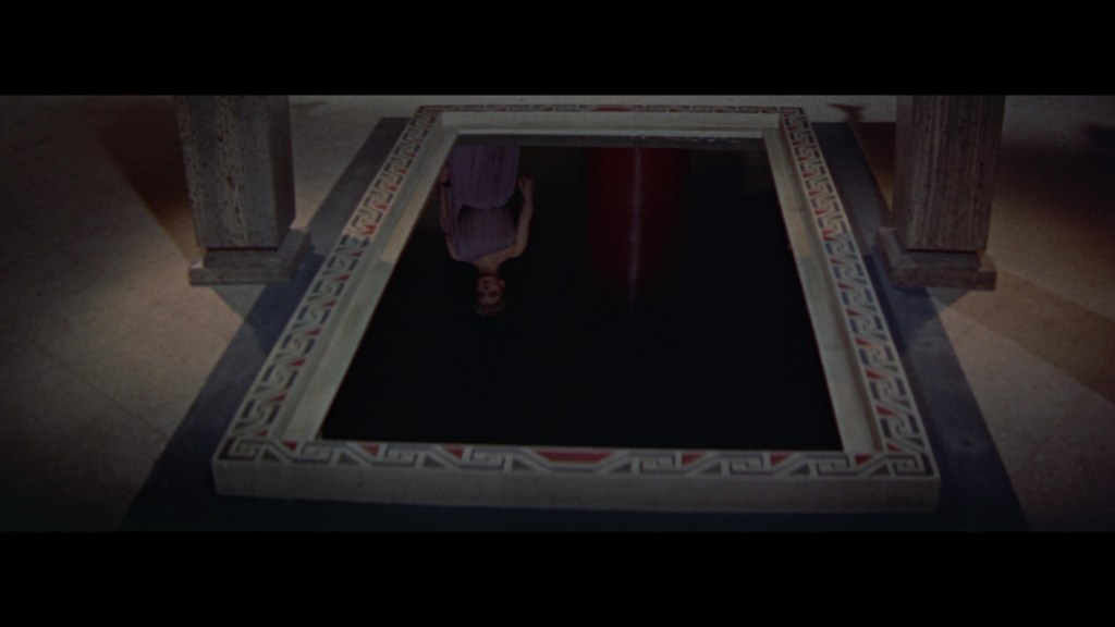 A dark, reflective pool framed by intricate patterns, with a faint reflection of a person in a flowing dress visible in the water.