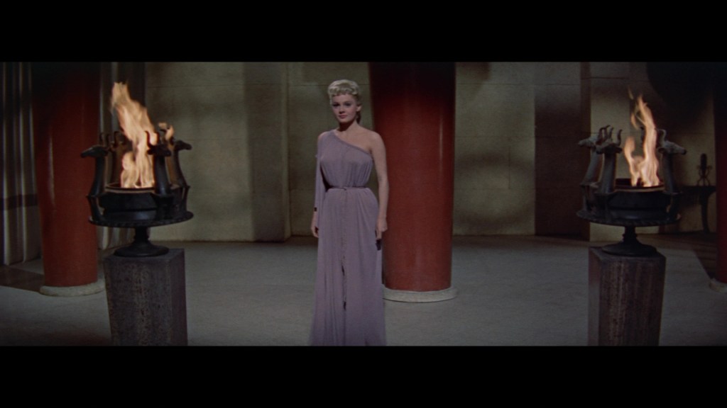A woman in a flowing lavender gown stands in a dimly lit room with two flaming torches on either side.