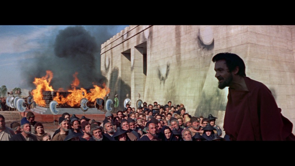 A crowd of people watching a speaker in a red robe, with a large fire burning behind them and a stone building in the background.