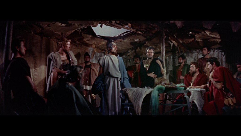 A dramatic scene featuring a group of men in ancient Roman attire, gathered around a table in a rustic setting. The man in the center, wearing a gray robe, appears to be addressing the group, who are dressed in various colored togas and cloaks, some with armor.