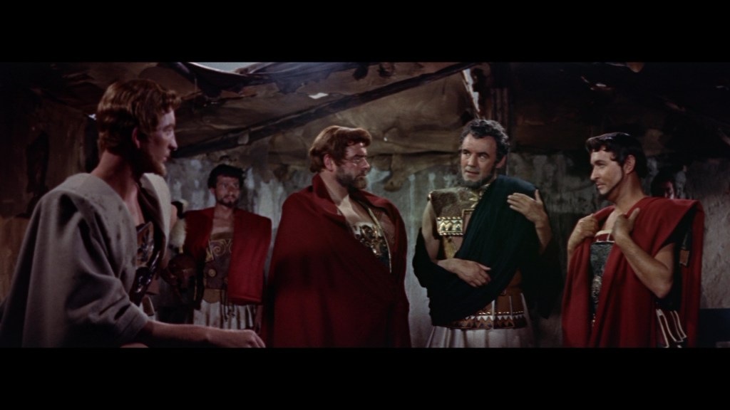 A group of five men dressed in ancient Roman attire, engaged in conversation, with expressions of concern and intrigue, set in a dimly lit indoor environment.