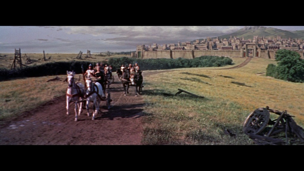 A group of horse riders approaches a large fortified city, set against a backdrop of rolling hills and a partly cloudy sky, with a grassy landscape and remnants of a wooden structure in the foreground.