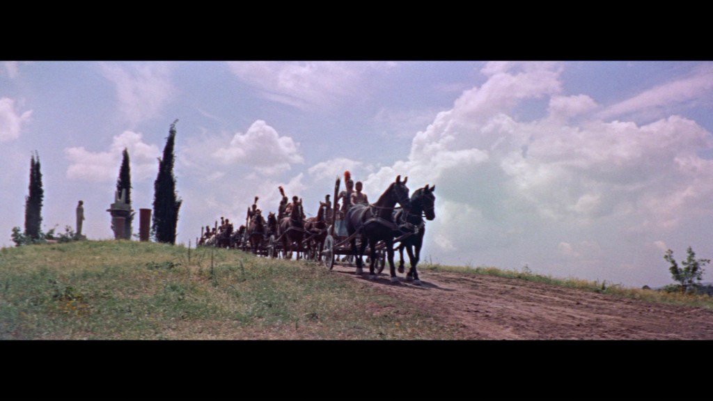 A procession of horses and riders on a dirt road, flanked by cypress trees, with a cloudy blue sky in the background.