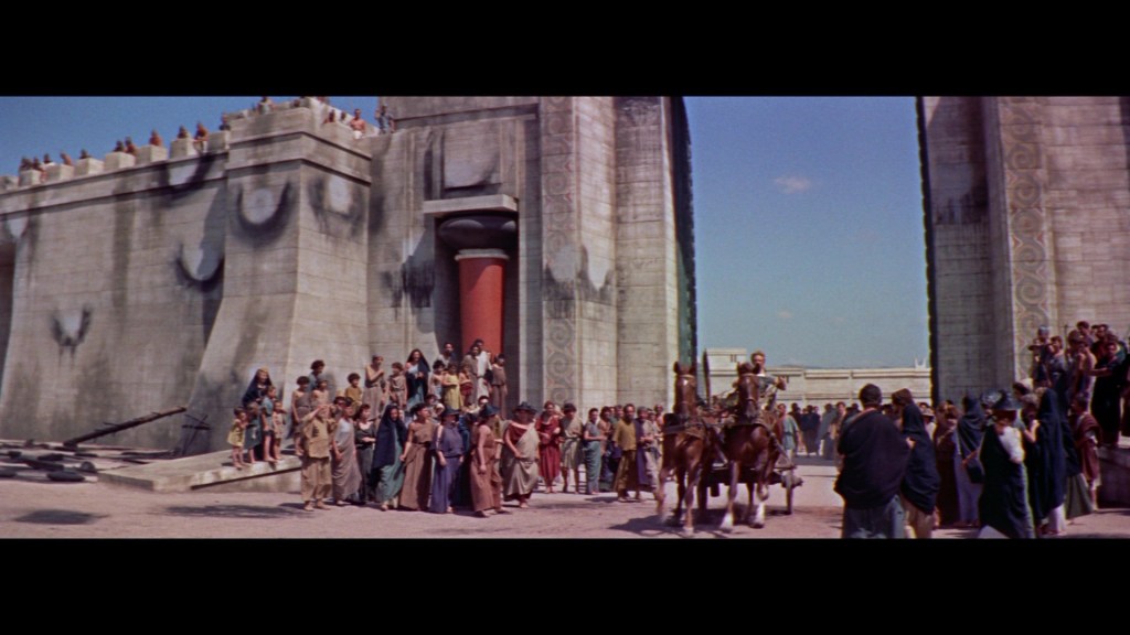 A historical scene depicting a large gathering of people in ancient attire outside a grand stone structure with ornate carvings, featuring a chariot pulled by two horses at the forefront.