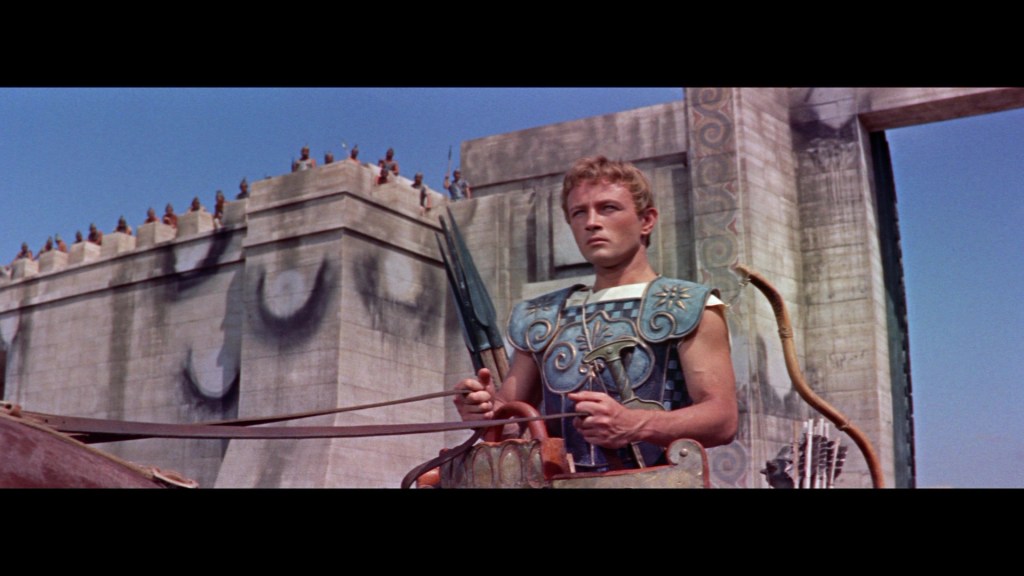 A young warrior dressed in ornate armor holds a bow and stands before a large stone structure, with spectators visible on the wall above.