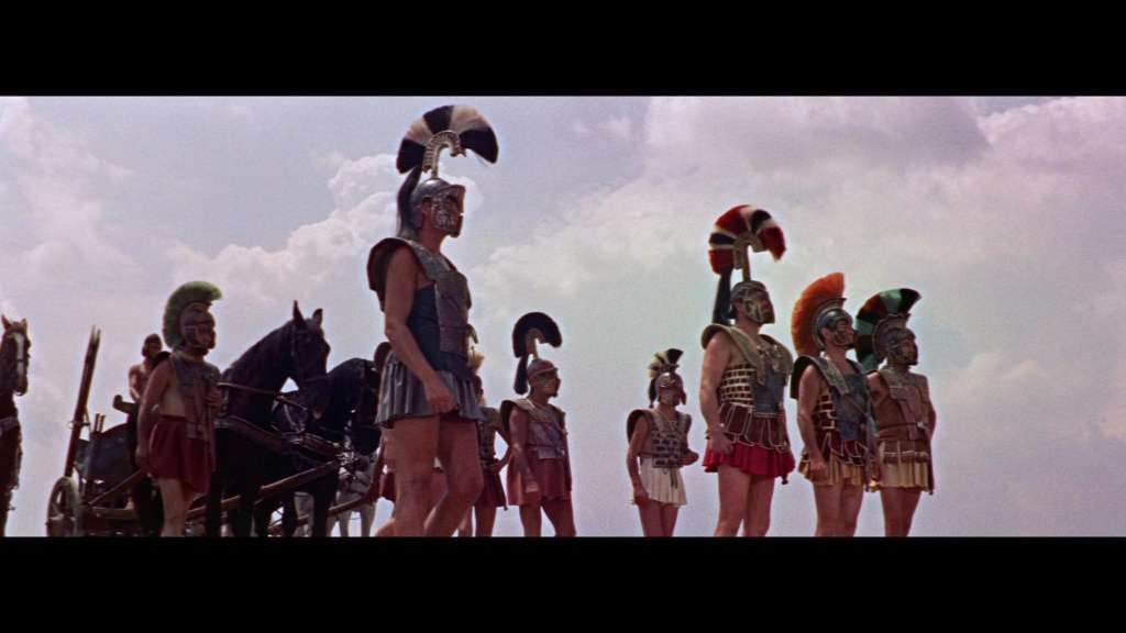 A group of ancient soldiers in armor and helmets, standing in formation against a cloudy sky, with a chariot and horse in the background.