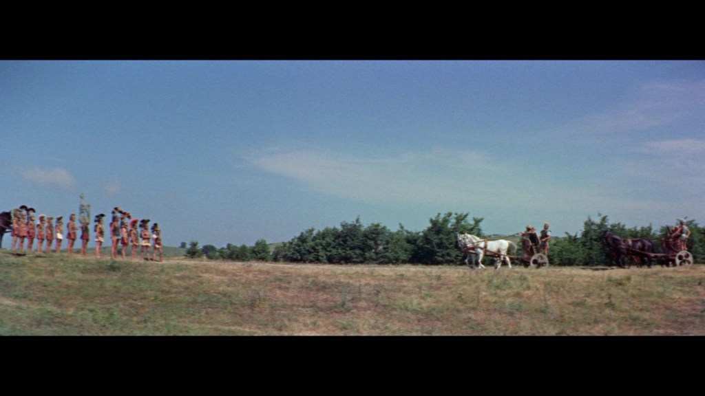 A group of people in traditional clothing gathered on a grassy field, facing a couple of horse-drawn carriages approaching from the right under a blue sky.