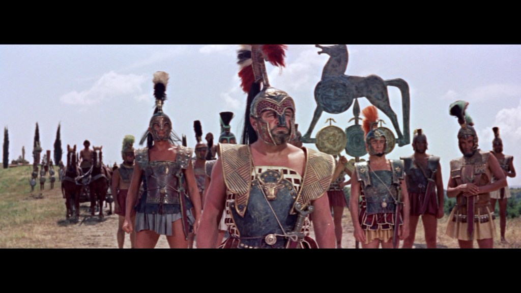A group of ancient warriors in armor, adorned with helmets and plumed crests, stand in formation on a grassy landscape under a blue sky. They are flanked by several horses, and a large bronze statue of a horse is visible in the background.