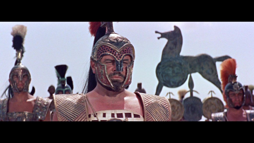 A group of armored warriors stands in a line, with a central figure wearing an elaborate helmet and breastplate. Behind them, a large wooden horse sculpture is visible against a clear sky.