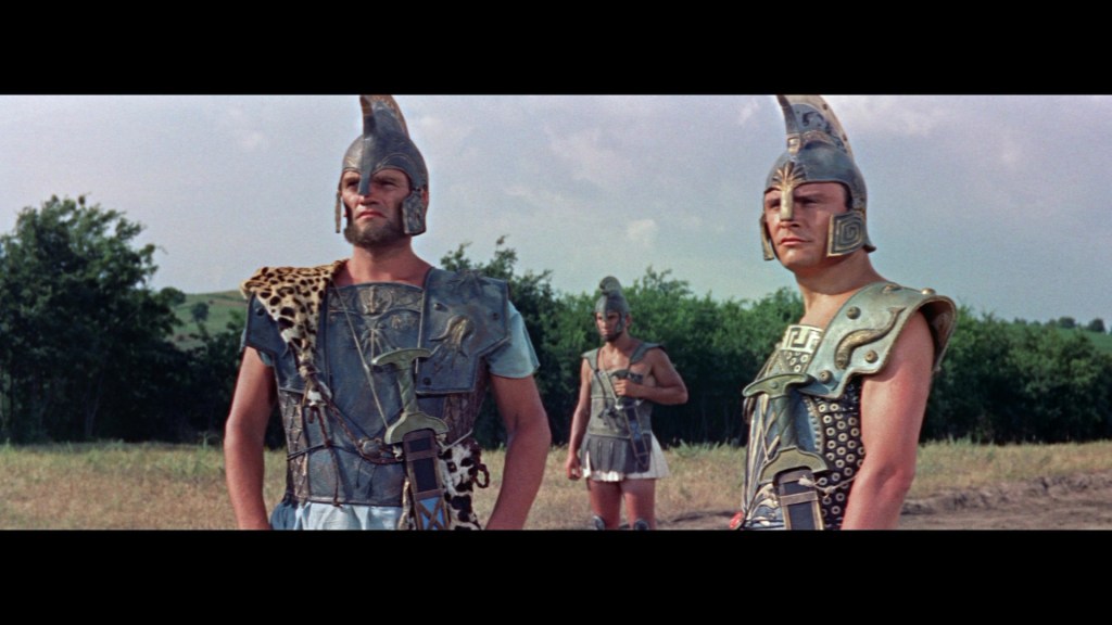 Three ancient warriors in elaborate armor and helmets stand in a grassy landscape, with trees in the background and a cloudy sky above.