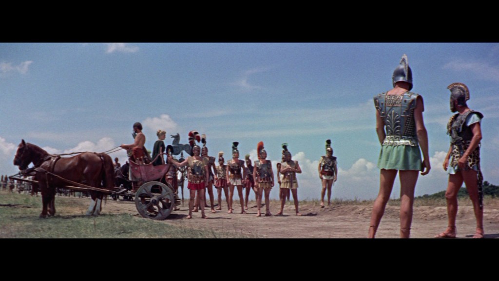 A scene depicting ancient warriors in armor, gathered in a field with a horse-drawn chariot. The sky is blue with some clouds.