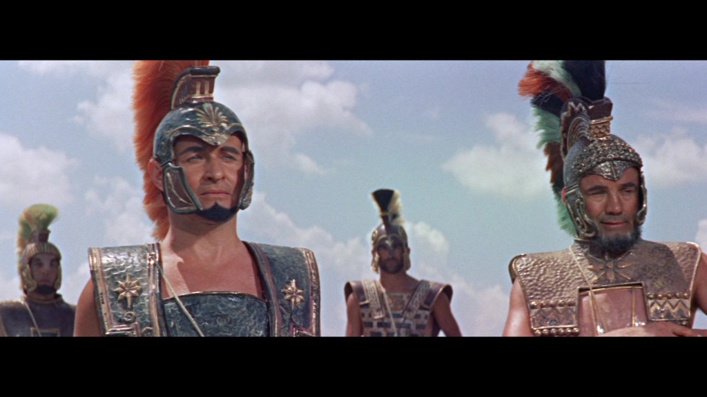 A group of ancient warriors in armor and helmets, standing against a blue sky with clouds. The foreground features two prominently dressed individuals, one with a red plume on their helmet, displaying an array of intricate armor details.