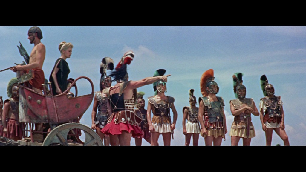 A scene depicting ancient warriors in armor, engaged in a tense confrontation, with one man pointing decisively, standing amidst a group of heavily armored soldiers and a woman in a chariot.