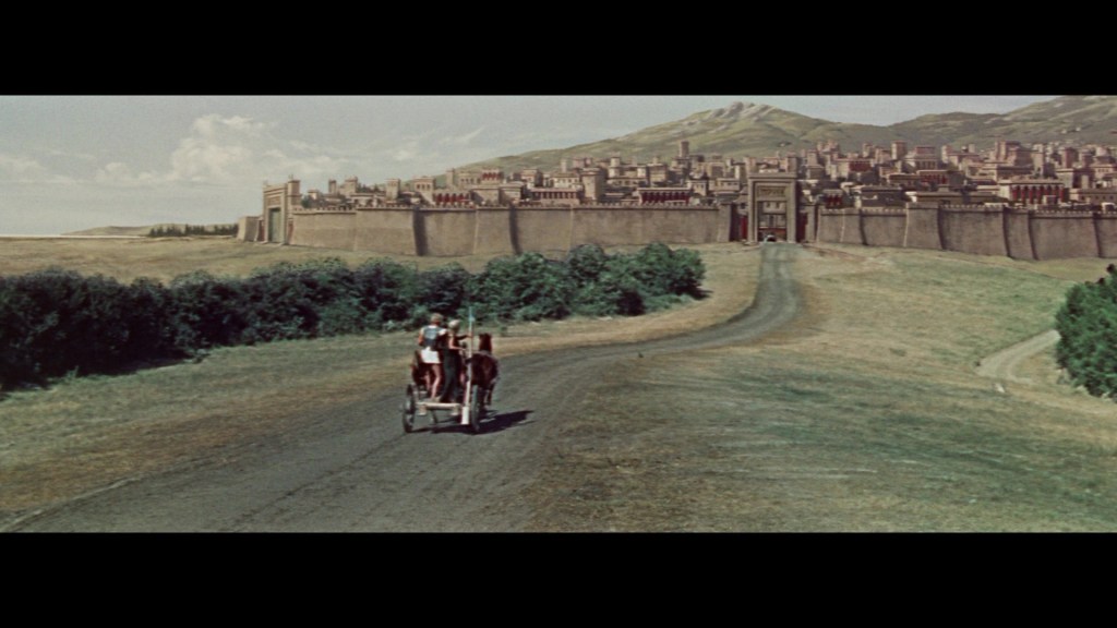 A scenic view of a dirt road leading towards a large ancient city with fortified walls, with two people riding in a horse-drawn cart in the foreground.