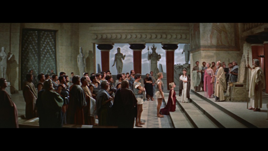A dramatic scene set in a classical Greek-style atrium, featuring a crowd of people dressed in ancient attire, gathered to witness a central event or speech. Statues and elaborate architectural details are visible in the background.