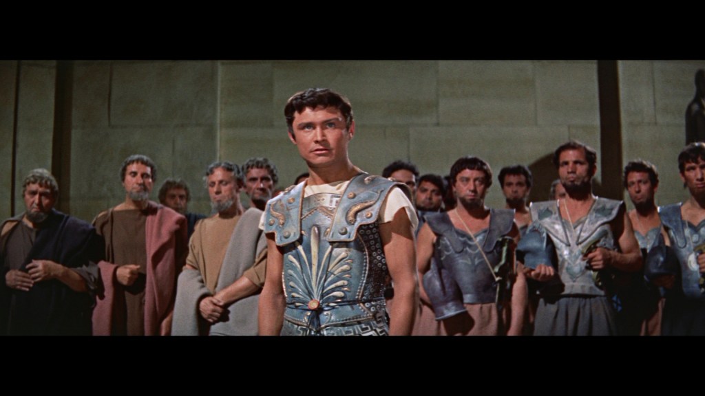 A young man in ancient armor stands at the forefront, looking determined, while a group of men in traditional attire surrounds him, conveying a sense of tension and anticipation in a historical setting.