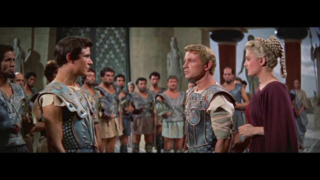 A scene depicting a tense conversation among soldiers in ancient Greek attire, with a prominent man in armor facing another man and a woman beside them, set in a grand architectural environment.
