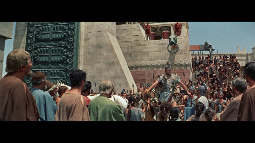 A crowd of people gathered in ancient attire watches a heroic figure in armor standing proudly on steps, surrounded by elaborate architecture.