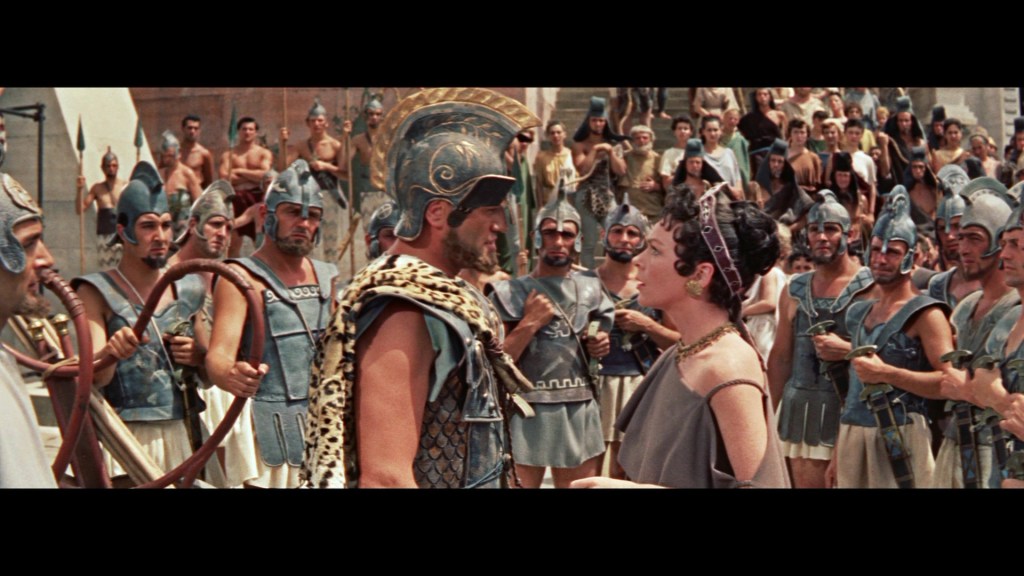 A dramatic scene depicting a confrontation between a male hero in ancient armor and a female character, surrounded by a crowd of soldiers and onlookers in a historical setting.