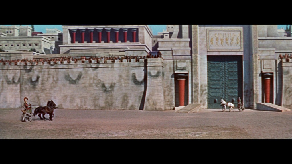 A scene depicting a large ancient city with a massive stone wall and ornate gates, where two horse-drawn chariots are present: one carrying a figure and the other empty, while a crowd watches from the top of the wall.