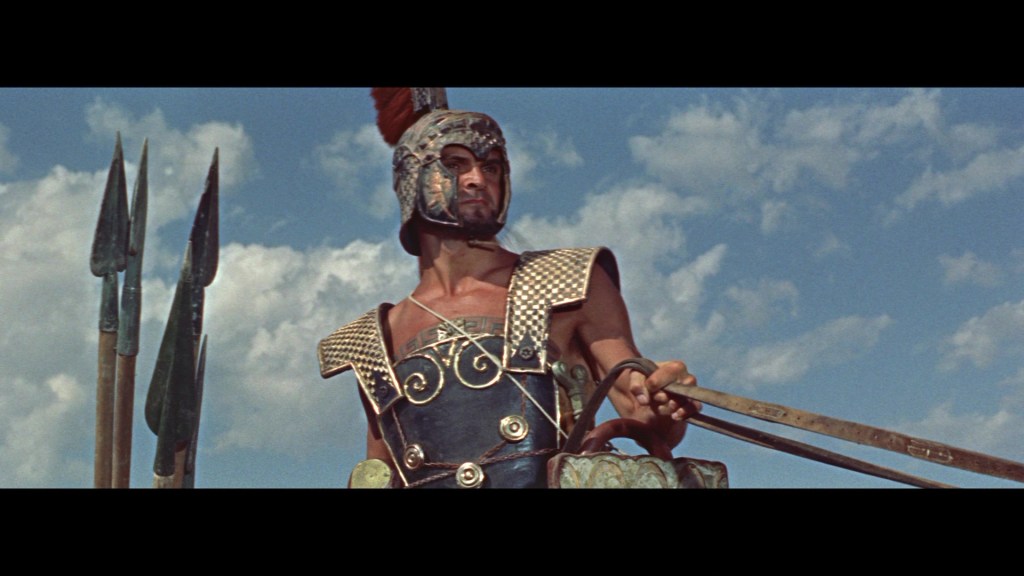 A muscular warrior in ancient armor and a helmet stands confidently, holding a sword with a backdrop of clouds and spear-like weapons.