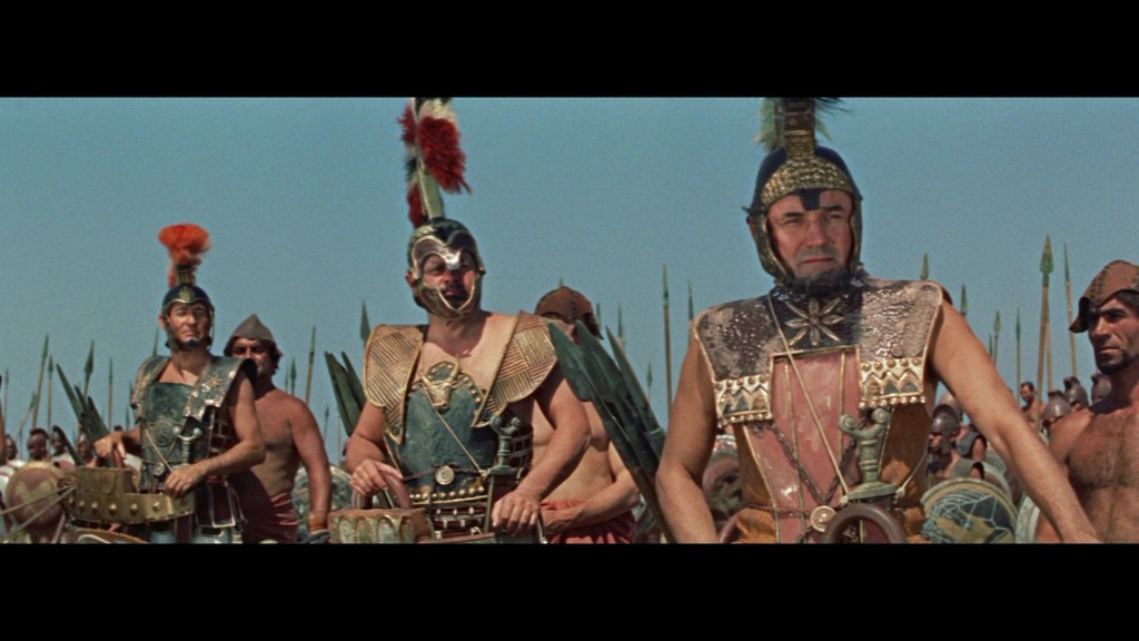 A scene depicting ancient warriors dressed in elaborate armor and helmets, standing in formation with weapons like spears, set against a clear blue sky.