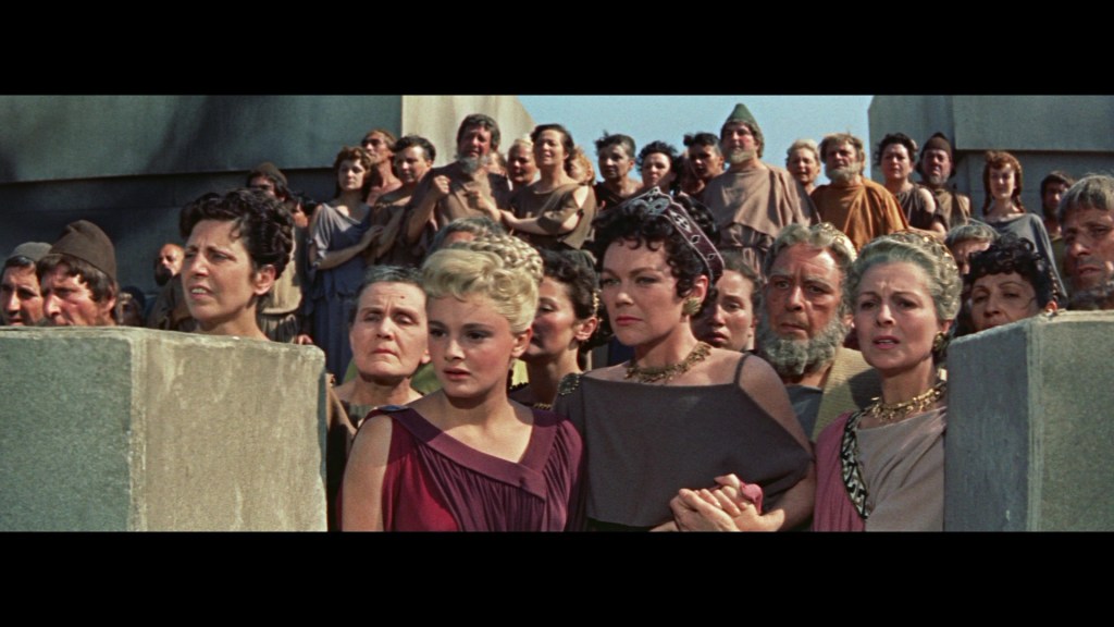 A crowd of people, both men and women, watches intently from behind a stone barrier. The foreground features two women holding hands, dressed in historical costumes, with expressions of concern. The background includes more spectators observing the scene.