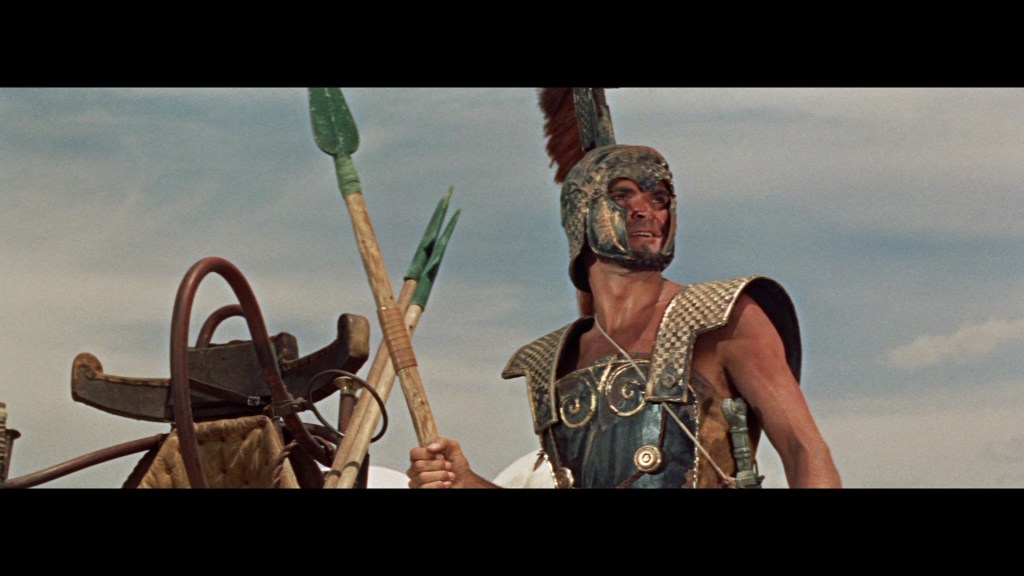 A warrior in ancient armor holds a spear, wearing a helmet with a feather plume, with a dramatic expression in a wide landscape under a cloudy sky.