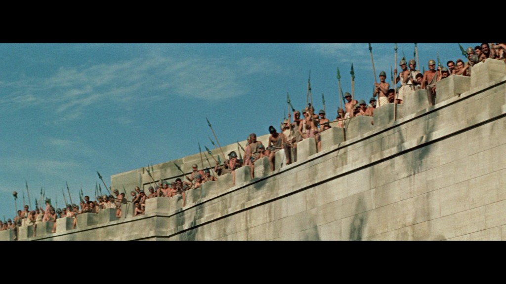A large group of people stands on the ramparts of a stone wall, holding spears, against a blue sky.
