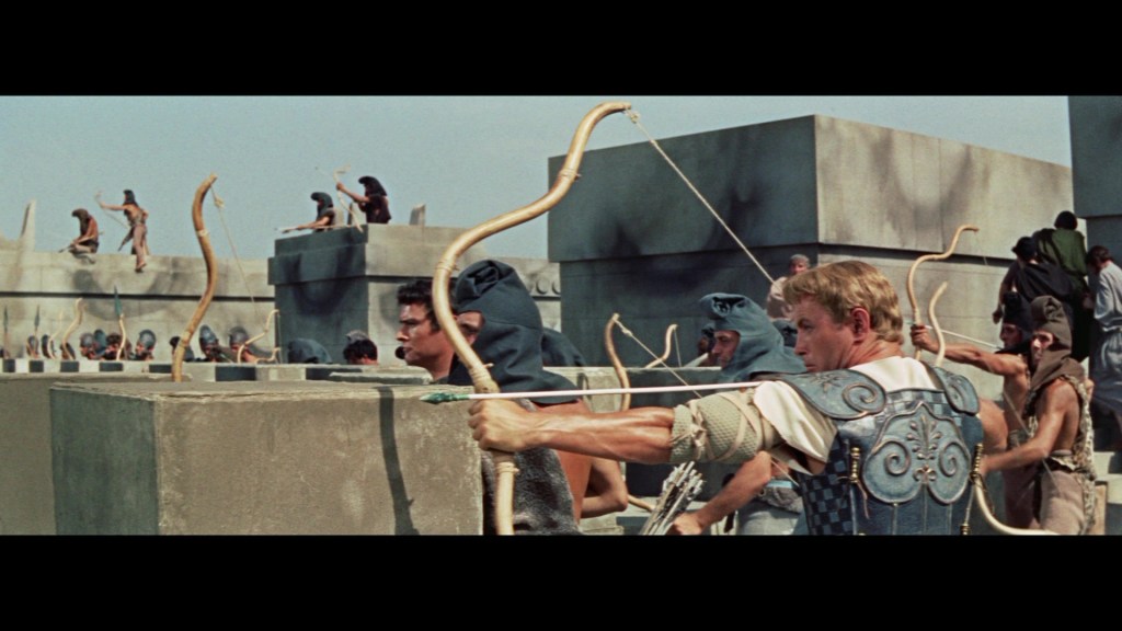 A scene depicting ancient warriors on a battleground, armed with bows and arrows, some aiming at an unseen target. They wear historical armor and costumes, set against a backdrop of stone walls and structures.