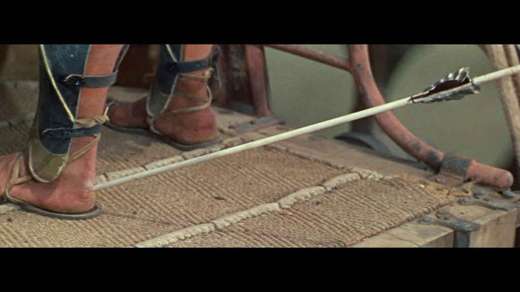 Close-up of a pair of feet wearing sandals, standing on a textured surface with a wooden background and an arrow resting nearby.