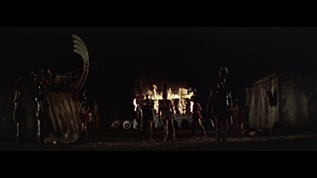 A nighttime scene depicting several warriors in ancient armor standing near a burning structure, with a distinct large statue or object in the background.