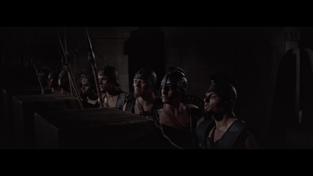 A group of ancient warriors in armor, standing in a darkened environment, preparing for battle with spears and shields.
