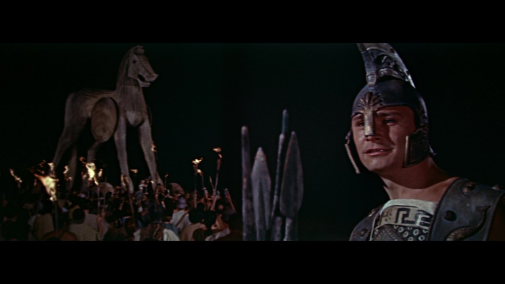 A scene featuring a large wooden horse with a crowd of people holding torches in the foreground, while a smiling warrior in armor stands in the foreground, gazing at the horse.