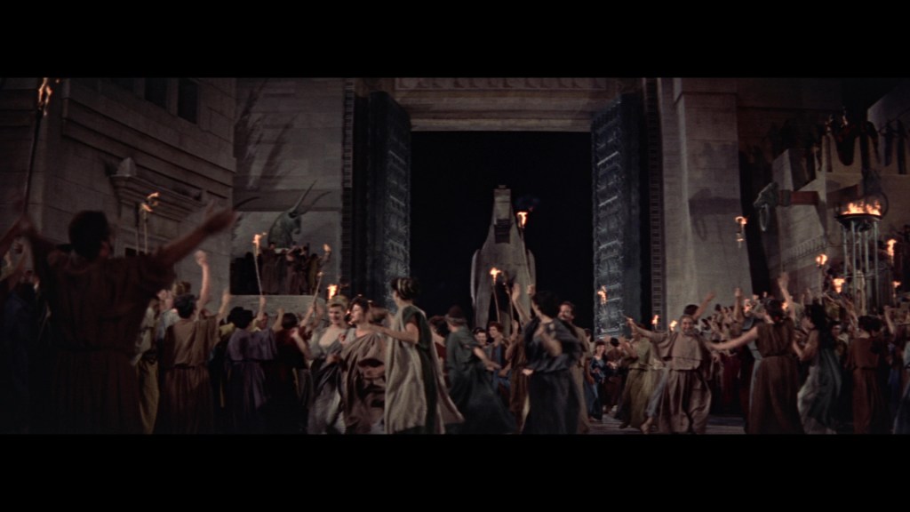 A large crowd of people dressed in ancient garments gathers at night, celebrating with torches in front of grand stone architecture and an imposing statue.