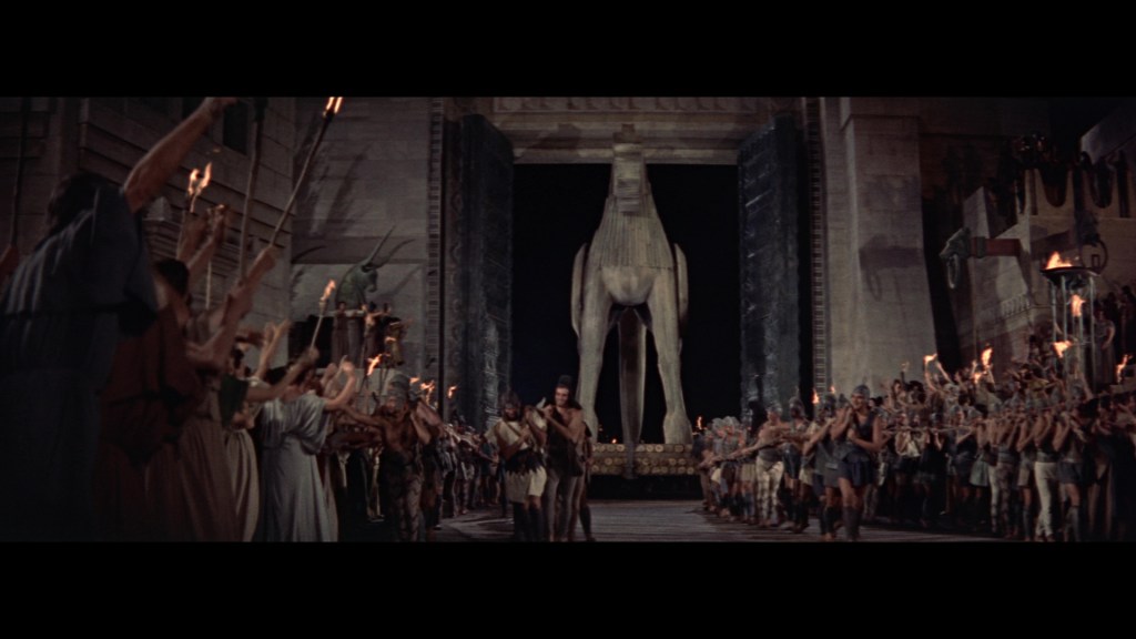 A large wooden horse is the focal point of the scene, surrounded by a crowd of people holding torches and weapons, celebrating in an ancient setting.