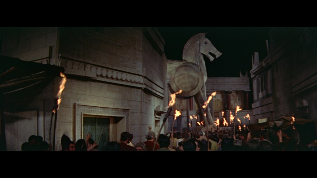 A nighttime scene featuring a large wooden horse statue, reminiscent of the Trojan Horse, surrounded by a crowd of people holding torches in an ancient city setting.