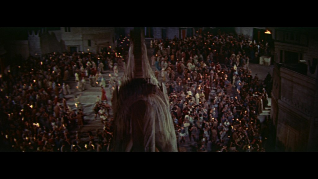 A large crowd of people gathered outdoors at night, holding torches in a bustling ancient setting with buildings in the background.