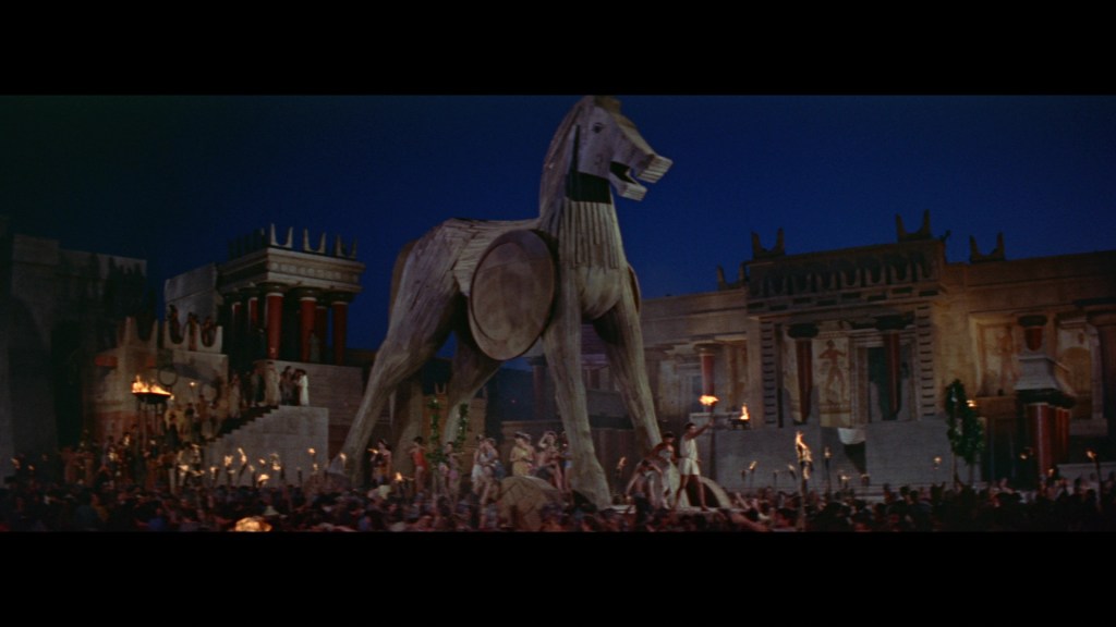 A large wooden horse statue stands in a bustling ancient city, surrounded by crowds of people holding torches, under a night sky.