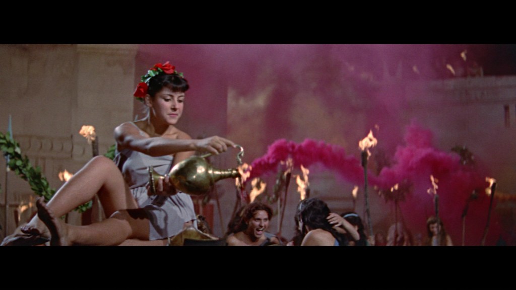 A woman in a flowing dress adorned with flowers sits on a ledge, pouring from a bronze jug. In the background, a vibrant scene unfolds with individuals celebrating around torches and clouds of pink smoke.
