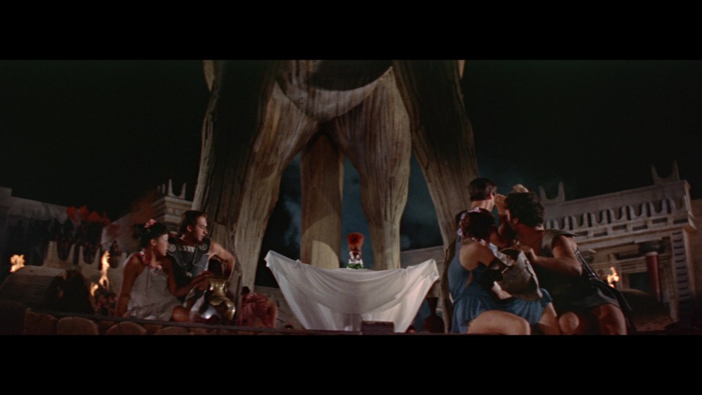 A dramatic scene featuring a large wooden statue in the background, with several characters seated in the foreground. The setting is illuminated by torches, suggesting a mysterious atmosphere.