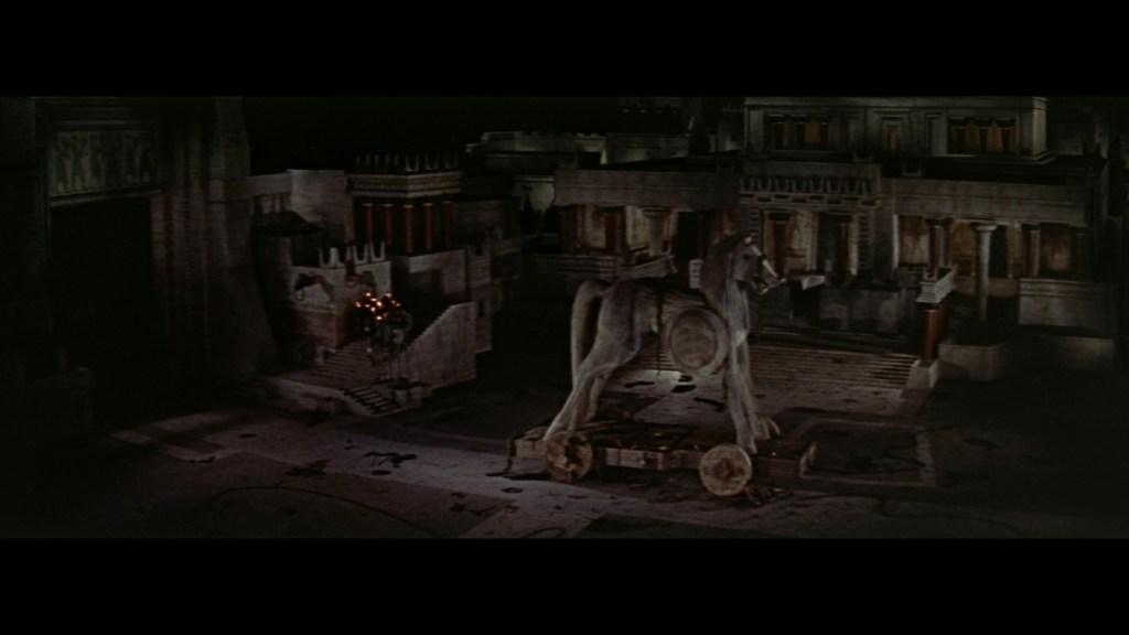A large wooden horse structure in a dimly lit ancient city setting, surrounded by ruins and towering buildings.
